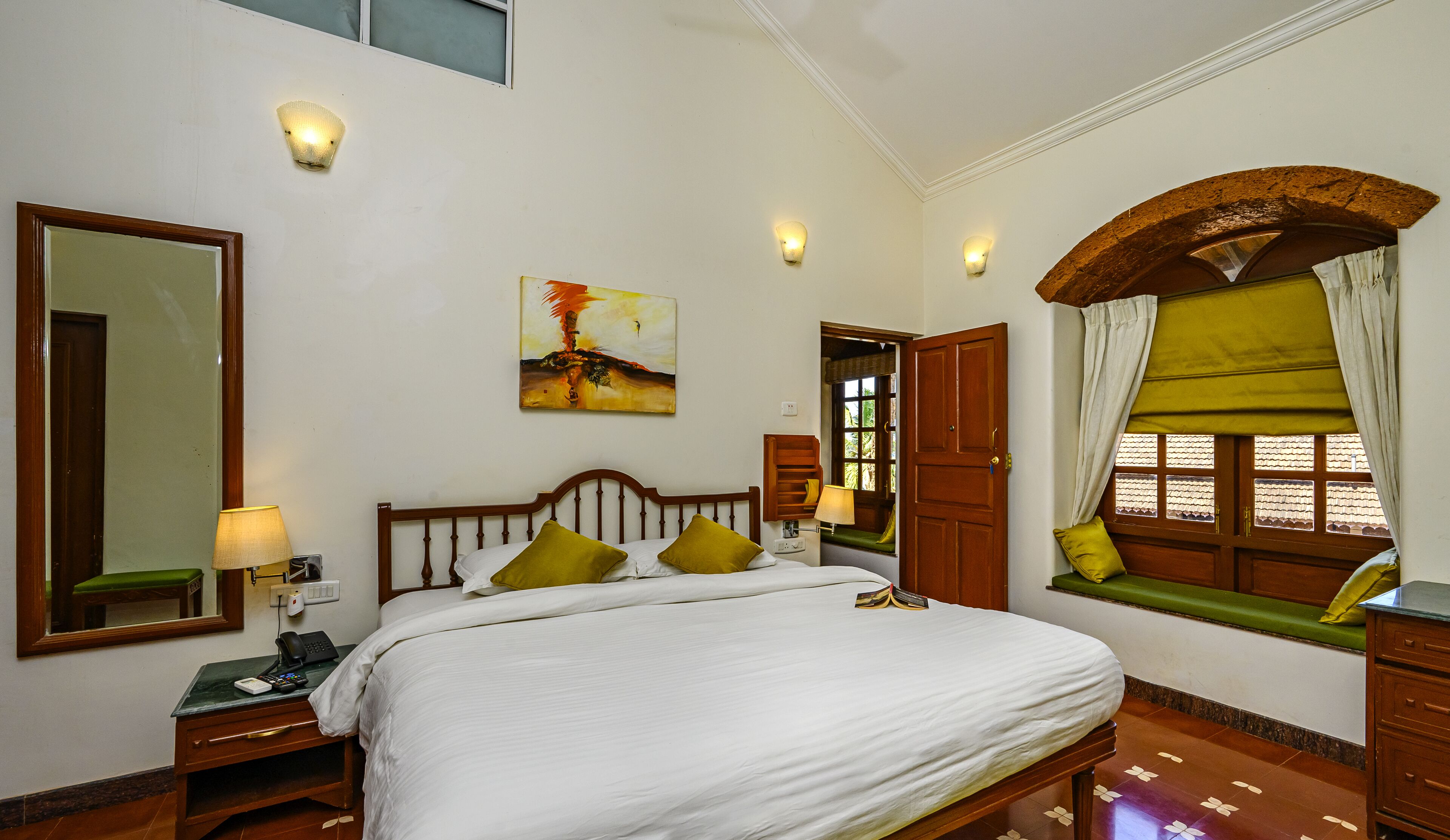 A hotel room with a bed, bedside lamps, a mirror, a painting, a window with curtains, and a wooden door.