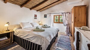 A bedroom with a large bed, two bedside tables with lamps, a wooden wardrobe, a window with curtains, and a small painting on the wall. - Rustic 3-bedroom Country Retreat With Pool, Free Wi-fi, and Breathtaking Views (Tavira)