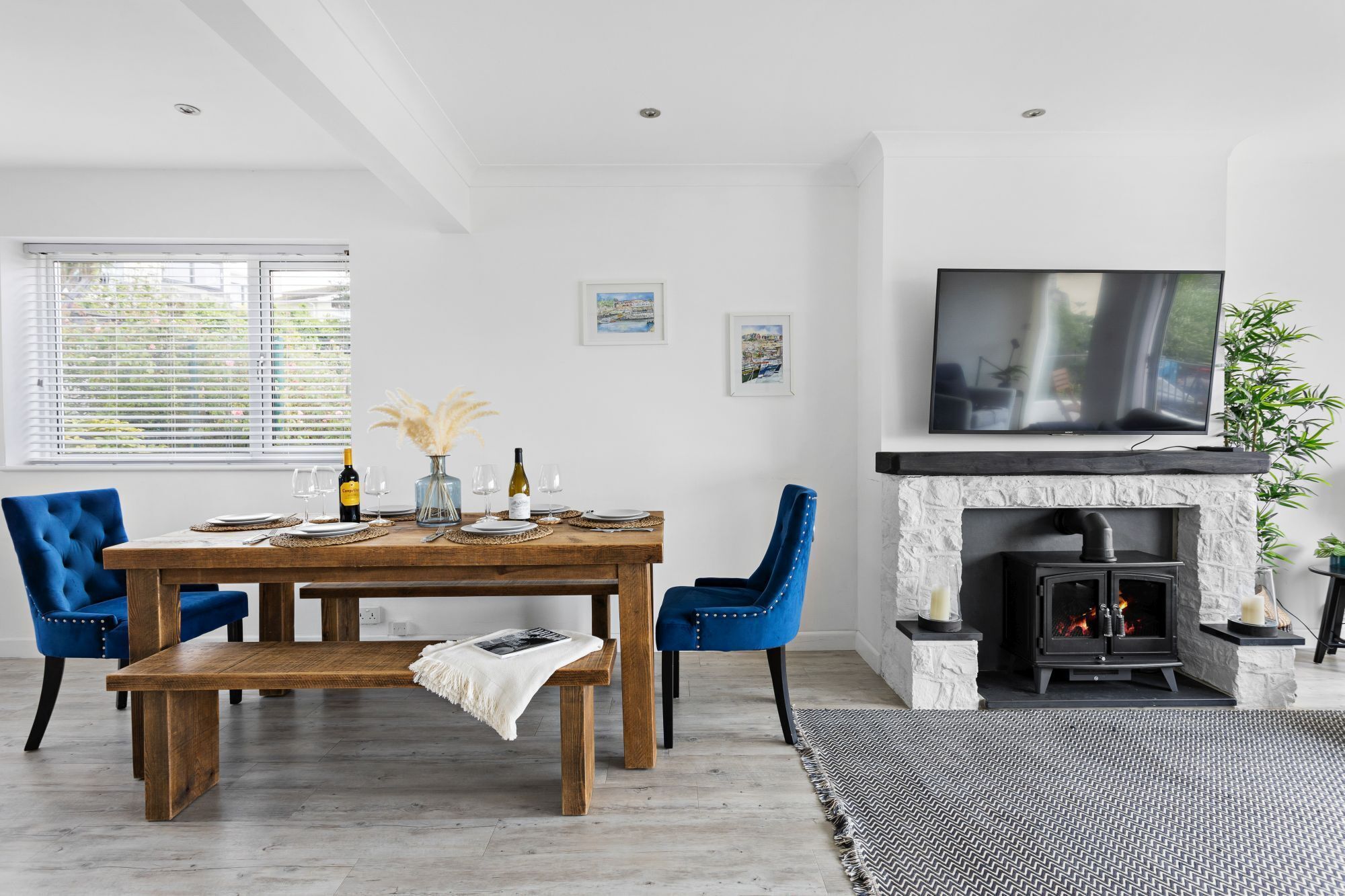 Een moderne woonkamer met een houten eettafel, blauwe stoelen, een open haard en een wandgemonteerde televisie.