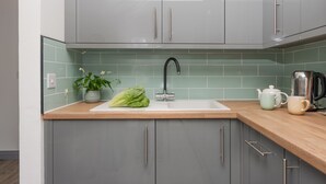 A modern kitchen with grey cabinets, a white sink, a green tiled backsplash, a potted plant, a cabbage, a kettle, and a mug on the countertop. - Hoopers Lodge,-Hot Tub, Pet Friendly (Crowlas)