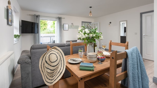 A cozy living room with a grey sofa, a wooden dining table set with books, glasses, and a vase of flowers. - Hoopers Lodge,-Hot Tub, Pet Friendly (Crowlas)