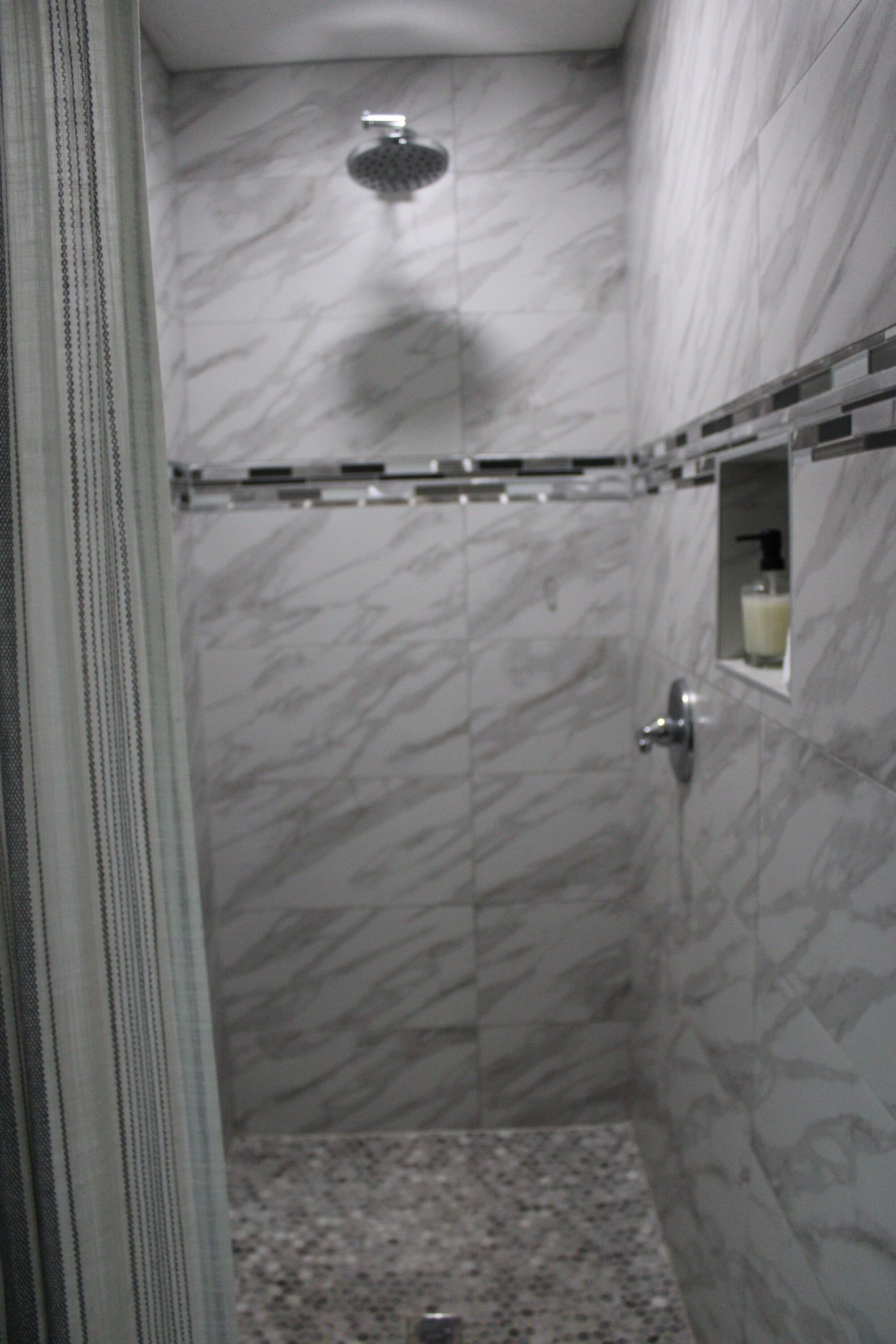 Un baño con paredes de mármol, ducha y un pequeño estante con una botella.