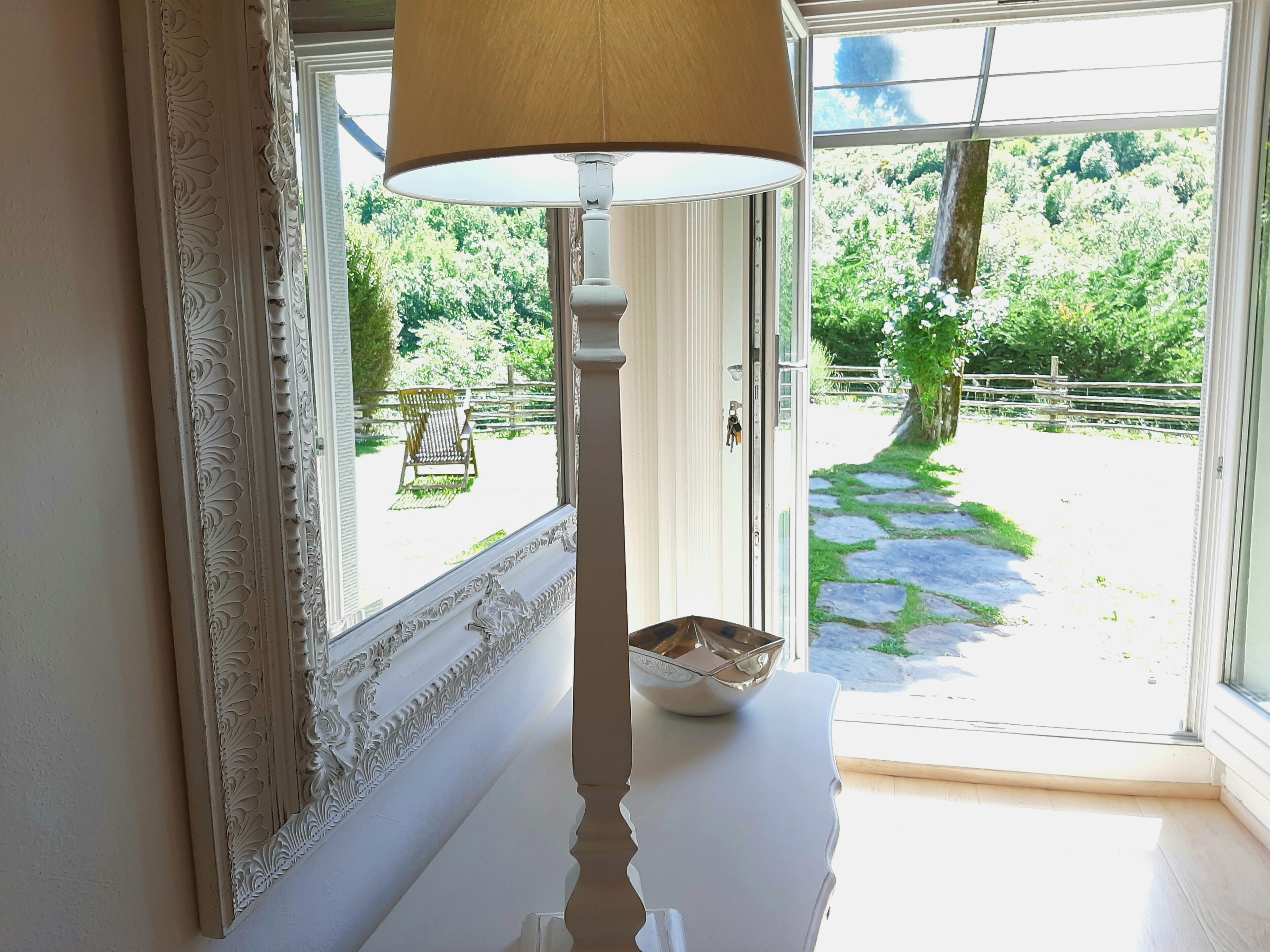 Een kamer met een witte, versierde spiegel, een witte tafel met een schaal, en uitzicht op een tuin door een glazen deur.