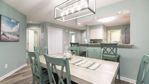 Un coin repas avec une table en bois et des chaises de couleur turquoise, une cuisine avec des meubles blancs et une suspension au-dessus de la table.