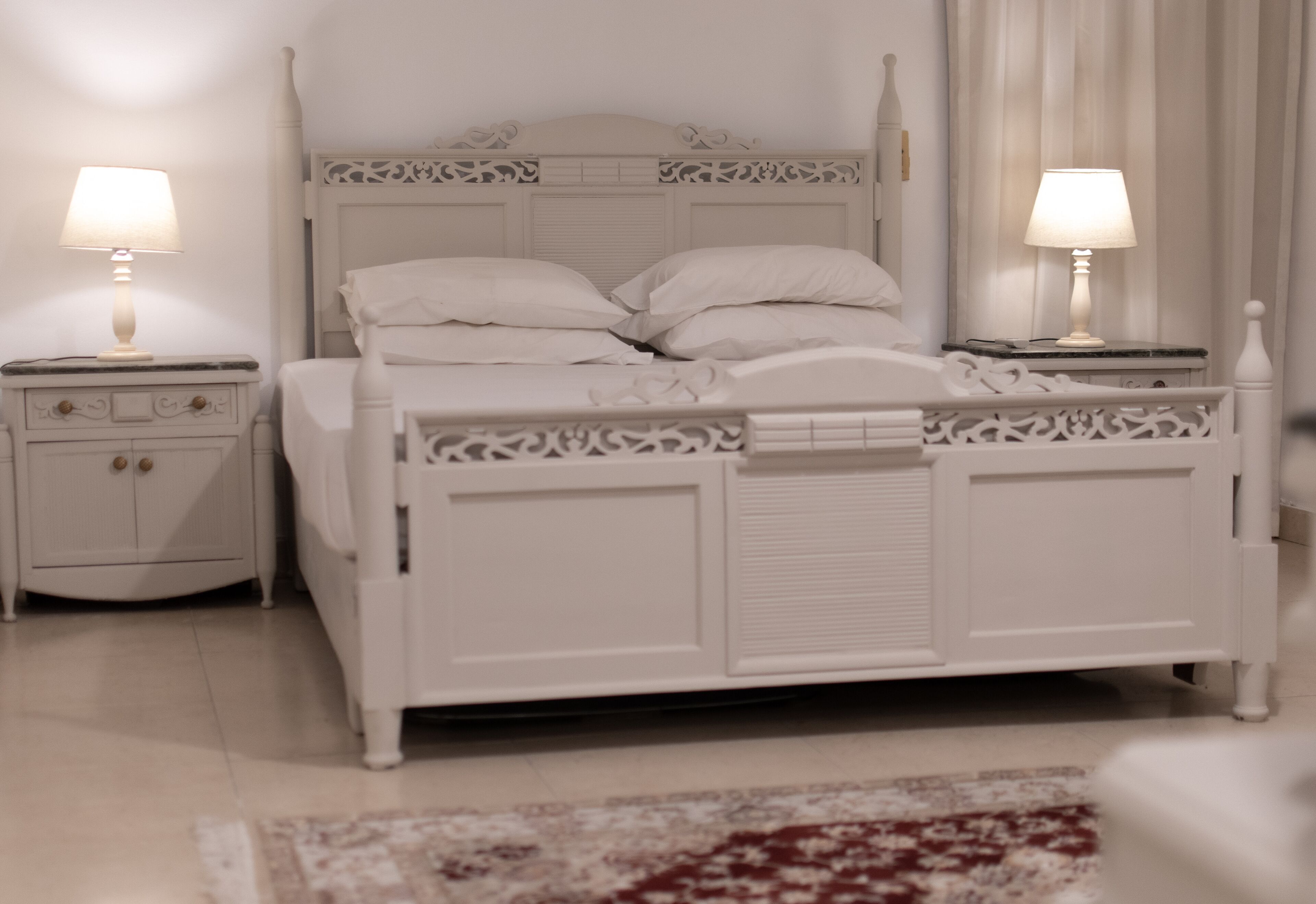 Ein Schlafzimmer mit einem weißen Bett, zwei Nachttischen mit Lampen, einem Beistelltisch und einem gemusterten Teppich.