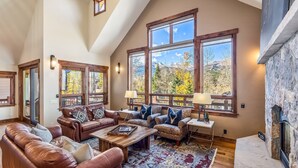 A spacious living room with leather sofas, a stone fireplace, large windows with mountain views, and a wooden ceiling. - BIG downtown home w/ views and great amenities! (Breckenridge)