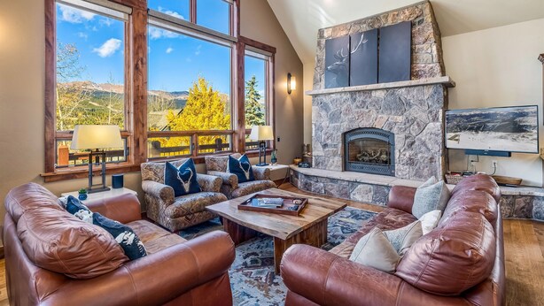 A cozy living room with leather sofas, a stone fireplace, and large windows offering a mountain view. - BIG downtown home w/ views and great amenities! (Breckenridge)
