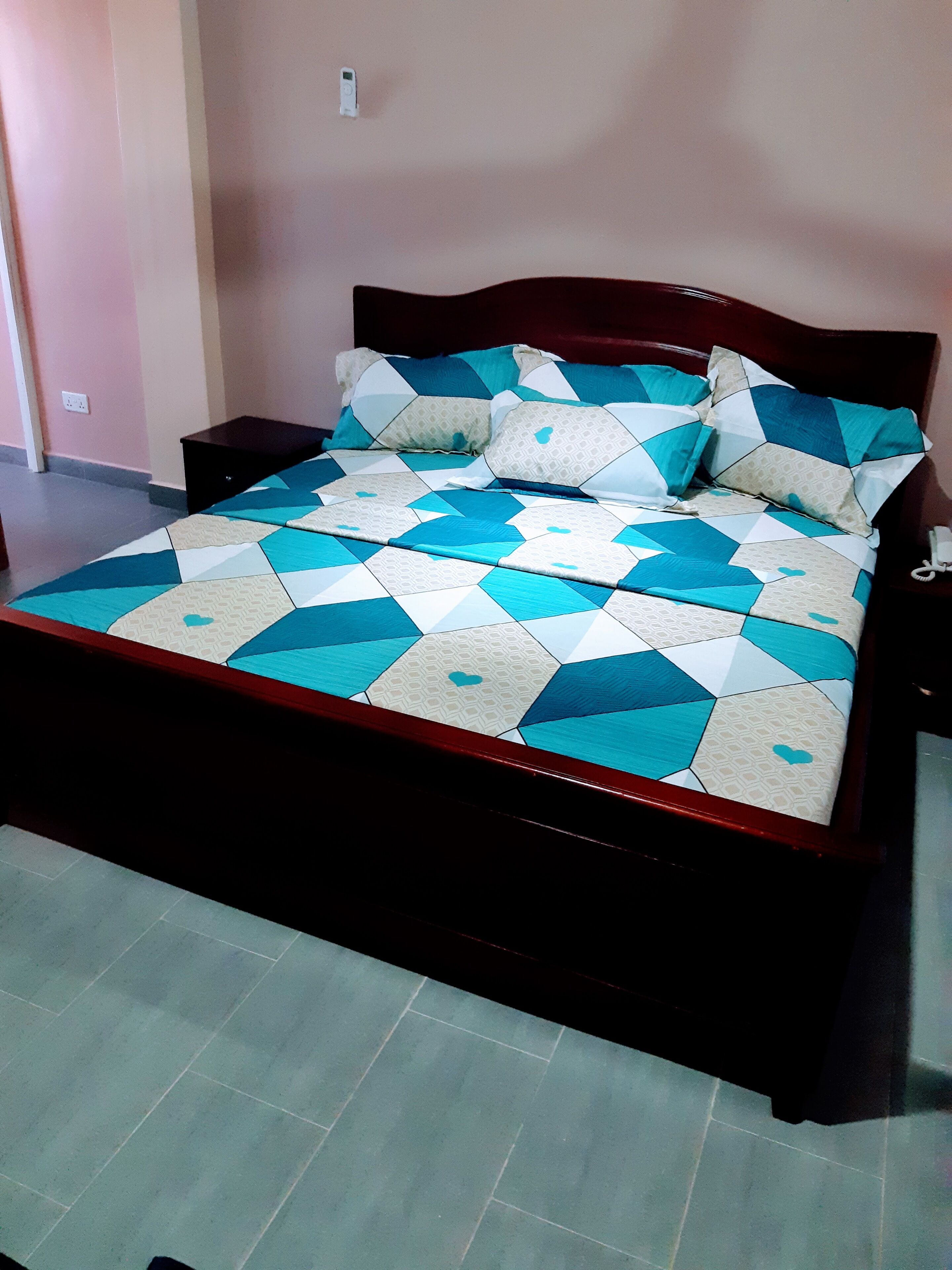 Un lit avec une couvre-lit à motifs géométriques et des coussins assortis, une tête de lit en bois, une table de chevet et une prise électrique murale.