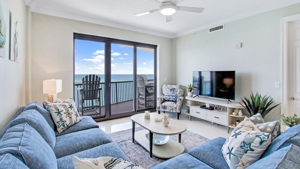 A living room with a large window, a ceiling fan, a flat-screen TV, and a sofa with decorative pillows. - Grand Pointe 708 by Liquid Life (Orange Beach)