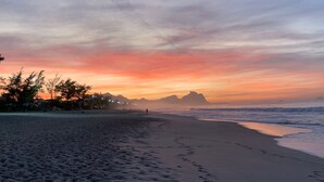 A beach with footprints leading to the water, a distant mountain, and a sunset sky.