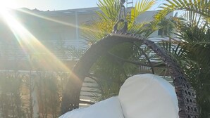 A wicker swing chair with white cushions on a wooden deck, overlooking a pool and palm trees.