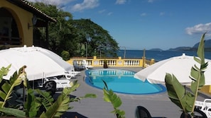 A pool area with white umbrellas and lounge chairs, surrounded by greenery and a view of the sea. - colonial hotel de ilhabela (Ilhabela)