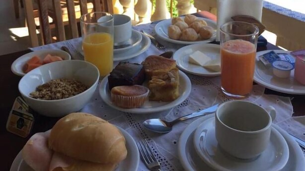 A breakfast table with plates of food, a glass of orange juice, and a view of a pool and mountains. - colonial hotel de ilhabela (Ilhabela)