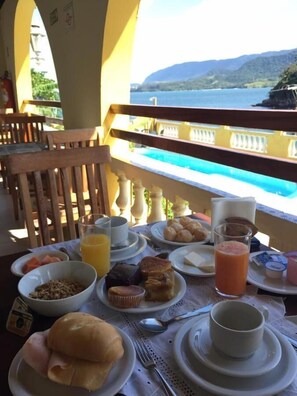 A breakfast table with plates of food, a glass of orange juice, and a view of a pool and mountains. - colonial hotel de ilhabela (Ilhabela)