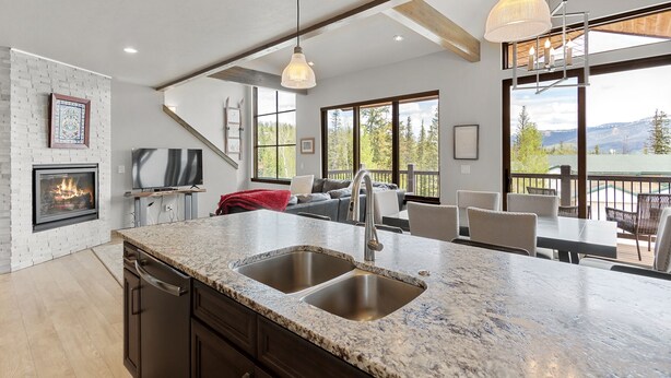 A modern kitchen with a large island, double sink, and a fireplace. There is a dining area with a view of the outdoors.