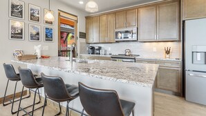 A modern kitchen with granite countertops, stainless steel appliances, and wooden cabinets. There are three bar stools and a large island with a sink.