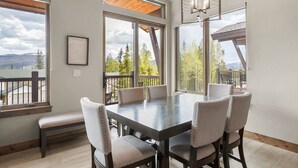 A dining area with a large table and six chairs, large windows with a view of trees and mountains, and a wooden ceiling.