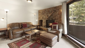 A cozy living room with a stone fireplace, leather sofa, ottoman, and a view of the outdoors. - Walk to the Marina and Amph in this central 2 BR Condo! (Dillon)