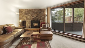 A living room with a stone fireplace, leather sofa, and a large window offering a view of buildings and trees. - Walk to the Marina and Amph in this central 2 BR Condo! (Dillon)
