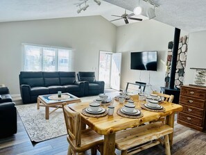 A spacious living room with a black leather sofa, a wooden dining table set with plates and glasses, a stone fireplace, and a TV mounted on the wall. - Cozy house in Indian River. Walking distance to Burt Lake and the public Beach. (Indian River)