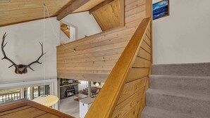 Uma escada de madeira leva a um mezanino com lareira, cabeça de veado montada na parede e vista para o exterior.
