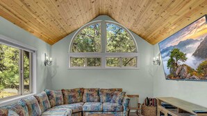 Uma sala de estar acolhedora com teto de madeira, um amplo sofá modular, lareira e uma grande janela com vista para árvores.