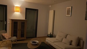 Een woonkamer met een bank, een voetenbankje en een kleine tafel met een kom. Er is een kast en een wandlamp.