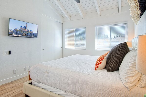A bedroom with a bed, a television mounted on the wall, and two windows with blinds.