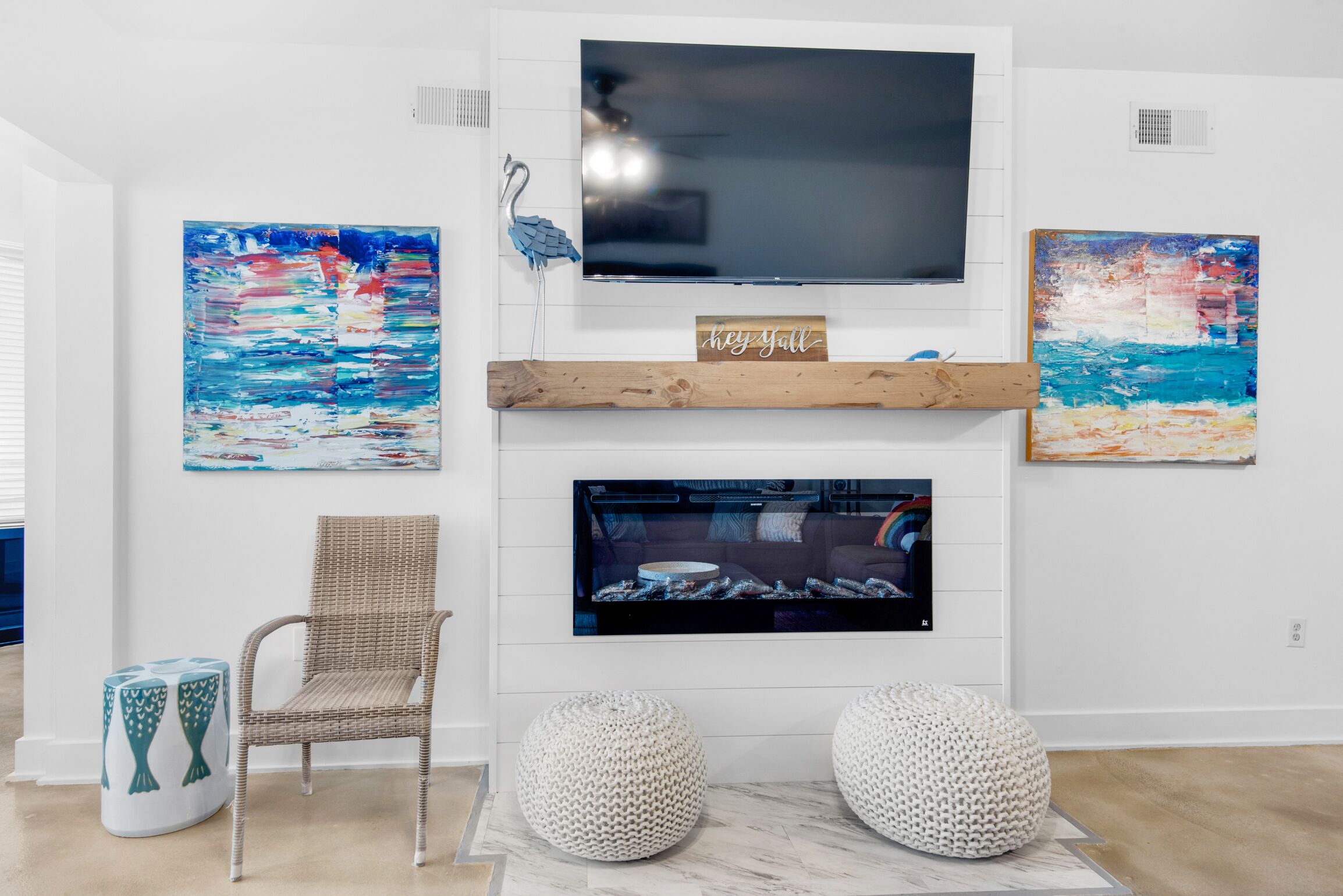 Un salon équipé d’un téléviseur à écran plat, de deux tableaux abstraits, d’un fauteuil en osier et de deux poufs ronds.