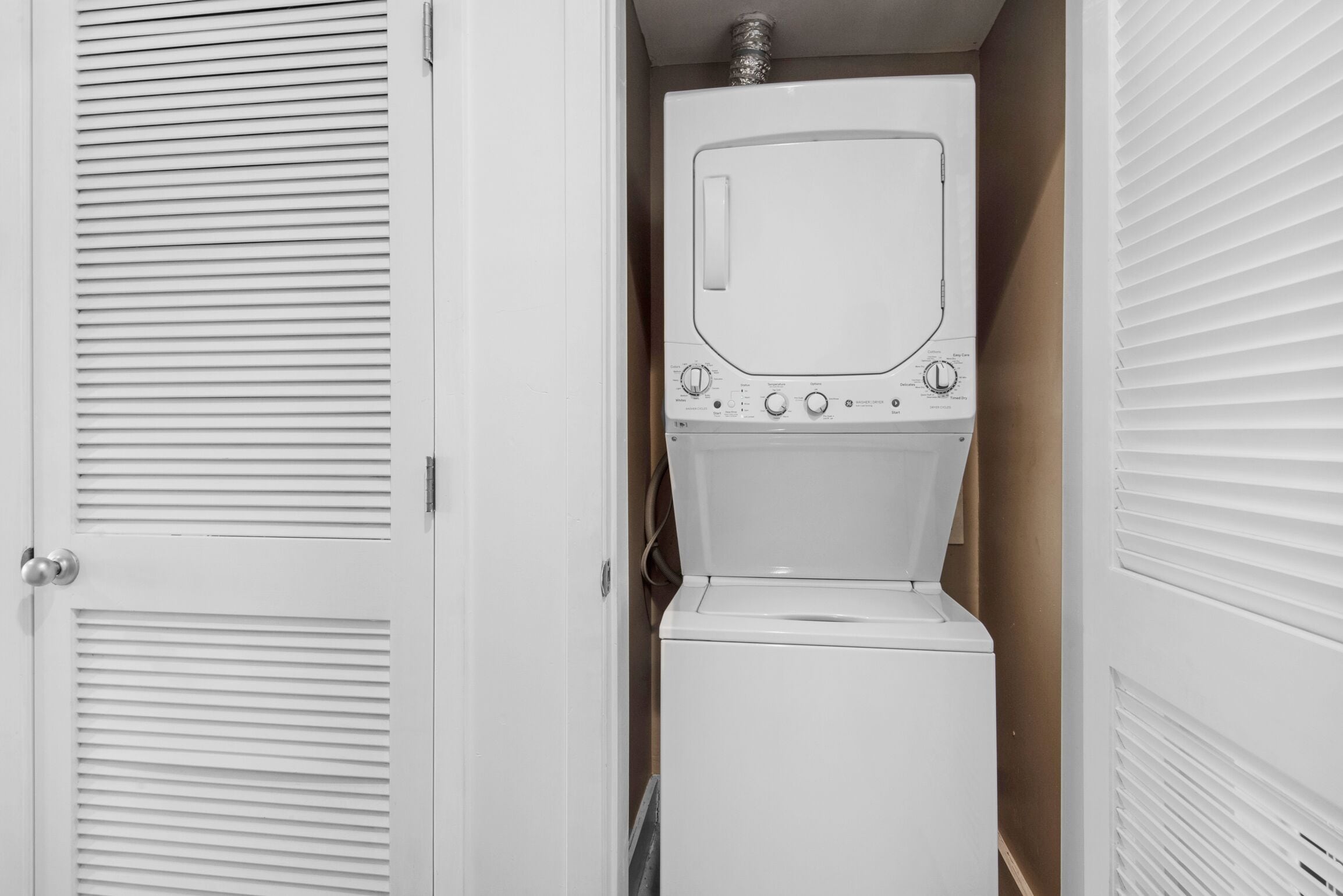 A compact laundry room with a washing machine and dryer, a door with louvered panels, and a vent on the ceiling.