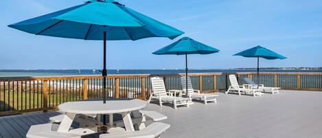 Une terrasse dotée de tables de pique-nique et de chaises longues blanches, de parasols bleus et offrant une vue sur l’océan.