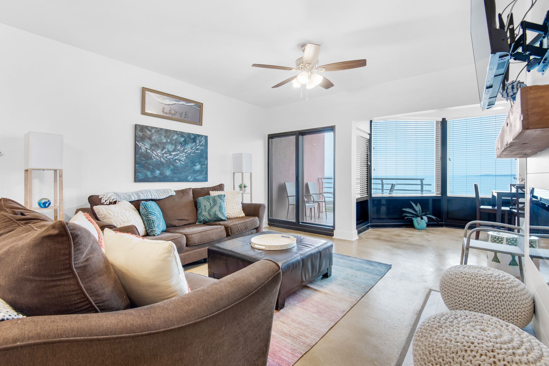 A modern living room with a brown sofa, a coffee table, and a ceiling fan. There is a large window with blinds and a view of the outdoors.
