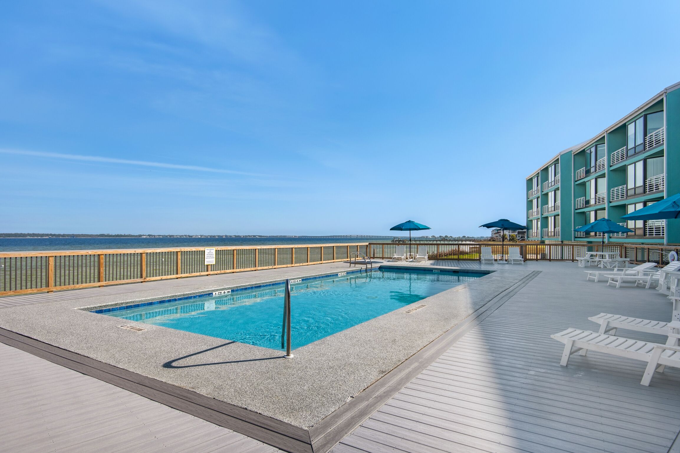 Une piscine rectangulaire entourée d’une terrasse en bois, de chaises longues et de parasols, le tout sur un balcon donnant sur l’océan.