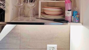 Eine Küche mit weißer Arbeitsplatte, einem Spülbecken und einem Regal mit Schüssel, Tellern und einer rosa Sprühflasche.