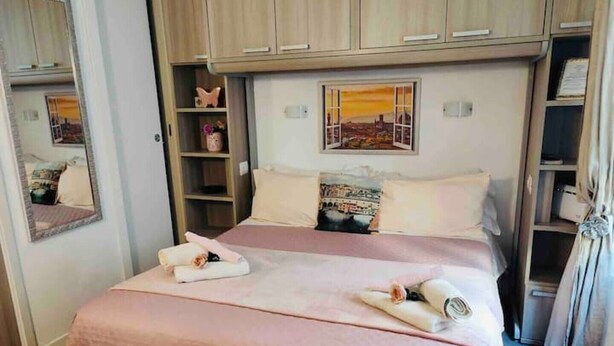 Ein ordentlich bezogenes Doppelbett mit rosa Bettwäsche, ein Nachttisch, ein Spiegel und ein Regal mit Dekoration.