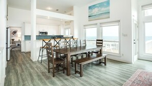A spacious dining area with a long wooden table and benches, a kitchen with white cabinets, and a view of the ocean through large windows. - Beachfront w/ Huge Deck, Dog Friendly, Dune Walkover, Feel Right at Home! (Galveston)
