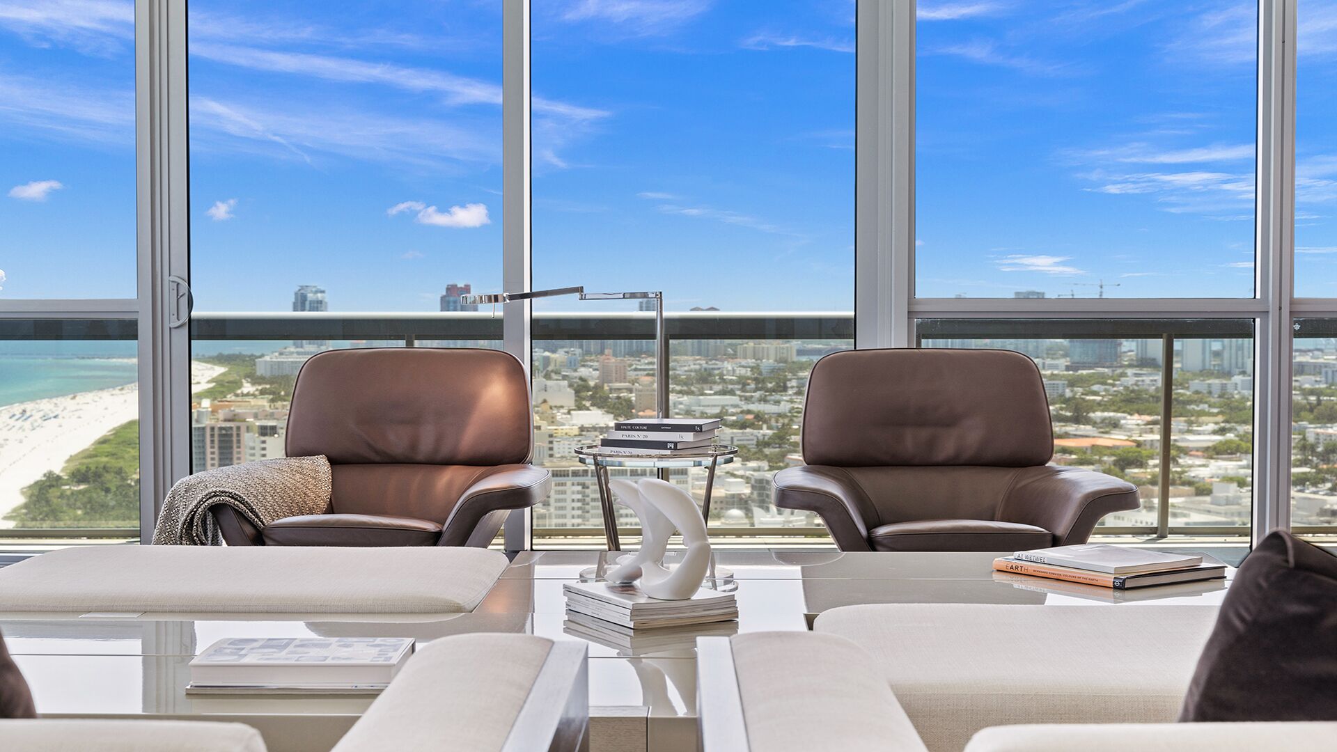 A modern office space with two leather chairs, a glass table, and a view of a cityscape and beach.