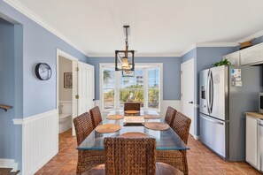 A dining area with a glass table and wicker chairs, a kitchen with a refrigerator, and a view through a window. - Community Pool, 3bdrm (Cape San Blas)
