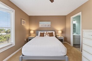 A bedroom with a large bed, two nightstands, a window with blinds, a painting on the wall, and a bathroom visible through an open door. - Community Pool, 3bdrm (Cape San Blas)