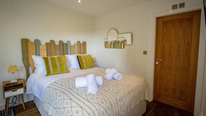 Een net opgemaakt bed met een geborduurde spreidek en een houten hoofdbord, en twee kussens. Er staat een nachtkastje met een lamp en een afstandsbediening. Achtergrond toont een houten deur.