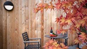Een houten terras met een ronde tafel en twee stoelen, een fles wijn en herfstbladeren.