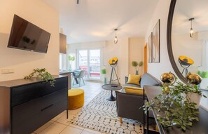 Un salon moderne avec un canapé, une table basse et un téléviseur fixé au mur. On y trouve également un coin repas avec des chaises et une table, ainsi qu’un grand miroir sur le mur.