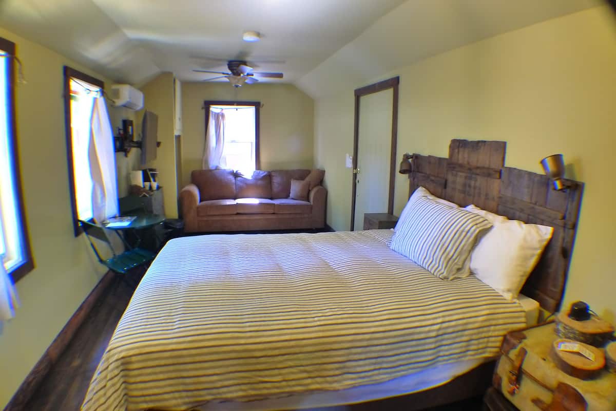 A bedroom with a large bed, a wooden headboard, a ceiling fan, a window with curtains, a small table, and a couch.