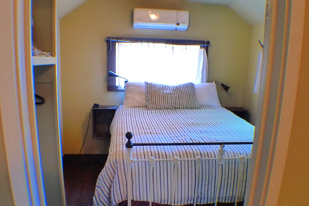 A narrow hallway leading to a bedroom with a bed, a nightstand, a ceiling fan, and a window with curtains.