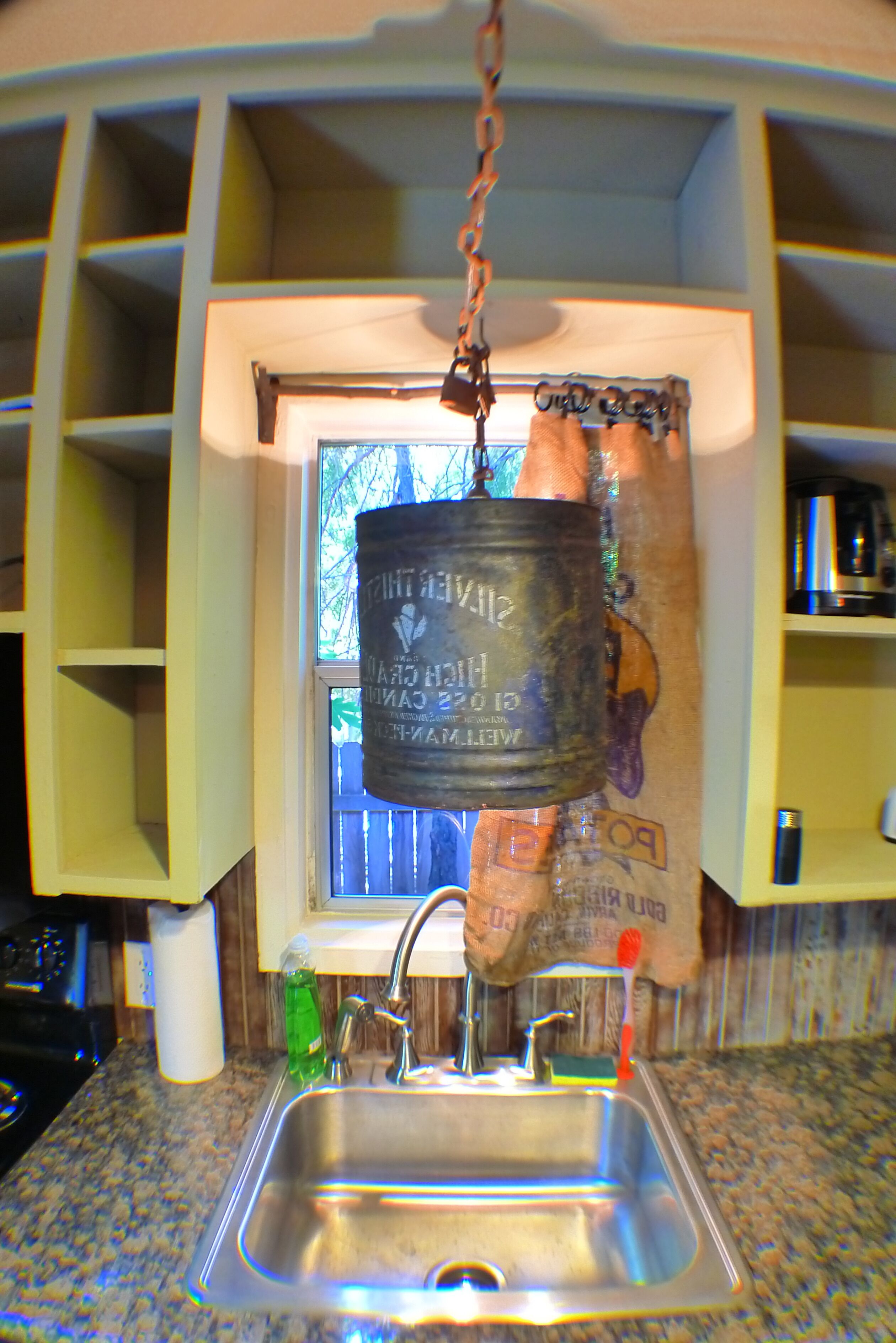 A kitchen with a stainless steel sink, a hanging pot rack, and a window with a view of trees.