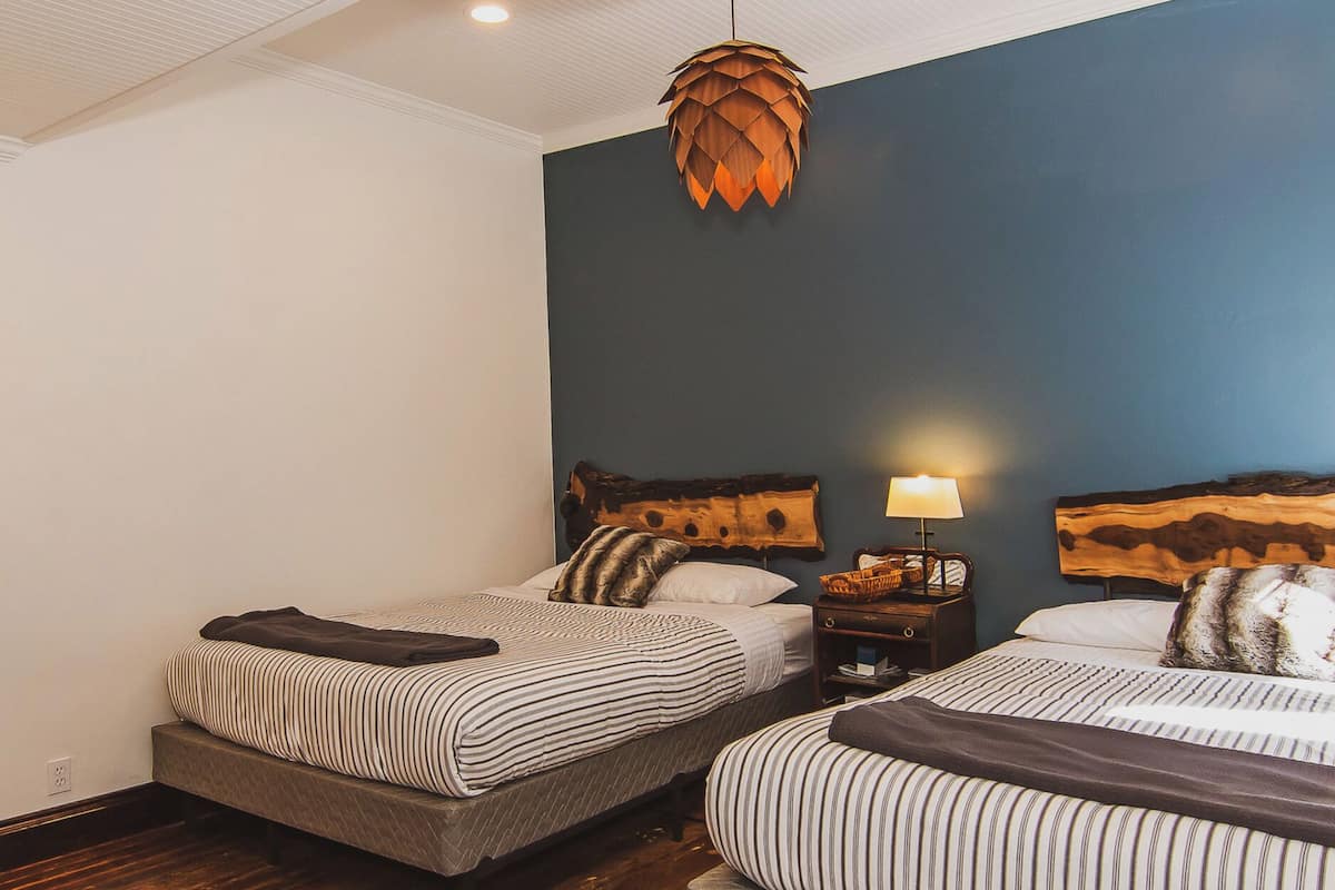 A hotel room with two beds, wooden flooring, a ceiling light, and a wall-mounted lamp.