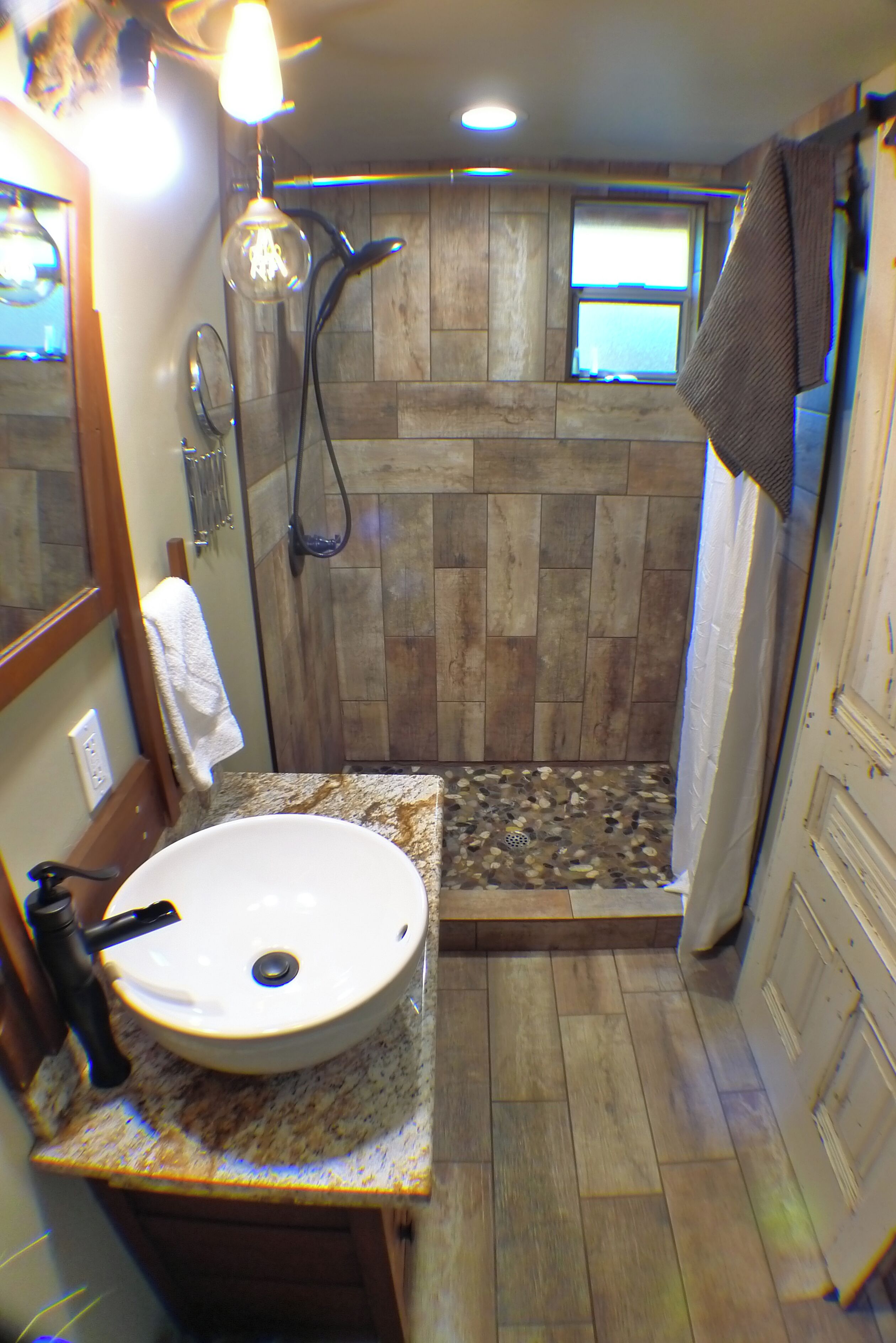 Une salle de bain avec un plancher en bois, un lavabo blanc, une douche avec une porte en bois et une serviette accrochée au mur.