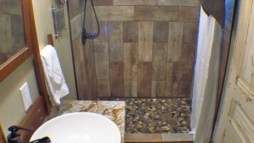 Une salle de bain avec un plancher en bois, un lavabo blanc, une douche avec une porte en bois et une serviette accrochée au mur.