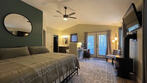 Une chambre à coucher comprenant un lit, un ventilateur de plafond, une télévision, un bureau avec une chaise, et une salle de bain avec un miroir et un lavabo.