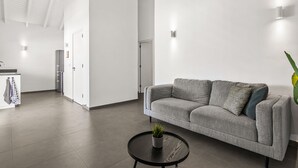 A modern living room with a grey sofa, a small round table with a plant, and a kitchen area in the background.